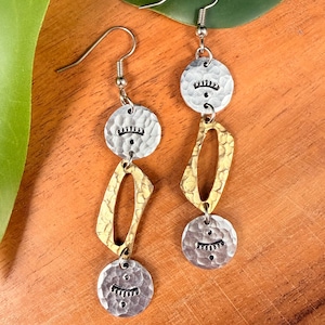 May include: Silver and gold-toned dangle earrings. Each earring features two hammered silver circles with a stamped eyelash design and a gold-toned, oblong shape. The earrings hang from silver ear wires and are displayed on a wooden surface.