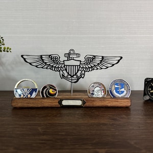 May include: A wooden display shelf with a metal US Navy Aviation badge and several challenge coins. The shelf has a small brass nameplate. A vintage clock is on the right. The coins have various designs and colors.