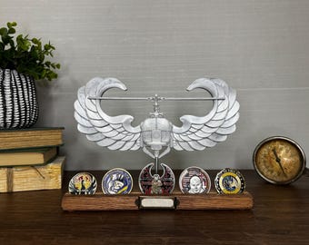 May include: A silver-colored metal helicopter rotor display stand with large wings, mounted on a rectangular wooden base. The stand holds five circular challenge coins in a row. A small blank metal plaque is centered on the front of the wooden base.