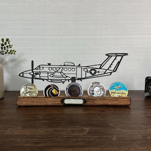 May include: A wooden display shelf with a black metal airplane silhouette and several collectible coins. The coins have various designs and colors. A small black clock is on the right side.