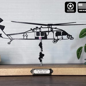 May include: Black metal helicopter silhouette mounted on a wooden base. The design includes two figures rappelling from the helicopter. A small plaque reads "O2 James Halpert 02 July 2025". The image includes the text "Veteran Owned Business" and "Made in USA".