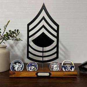 May include: A wooden display stand with four challenge coins, a black metal rank insignia, and a small compass. The coins have colorful designs and are displayed on a wooden base. A small ceramic vase with greenery is on the left.