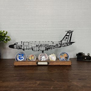 May include: A black metal airplane silhouette with "U.S. AIR FORCE" text sits above a wooden display holding five military challenge coins. The coins are round with colorful designs. A small black telegraph key is on the right.