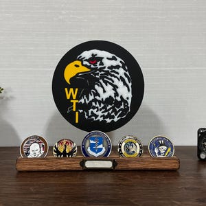 May include: A wooden display stand holding five military challenge coins, with a large black circular emblem of an eagle's head in the background. The coins feature various designs and colors. A small clock is on the right.