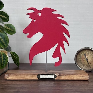 May include: A red horse head silhouette sculpture on a wooden base. The sculpture is mounted on a metal rod. A small metal tag with text is attached to the base. A vintage gauge is visible in the background.