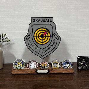 May include: A display featuring a gray and yellow US Air Force Weapons School Graduate emblem, a wooden coin holder with five military challenge coins, and a small black clock. The coins have colorful designs.