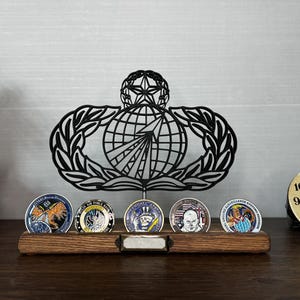 May include: A black metal United States Air Force Space Operations badge display stand featuring a globe and star emblem. The stand includes a wooden base with five circular challenge coins arranged in a row and a blank metal nameplate in the center.
