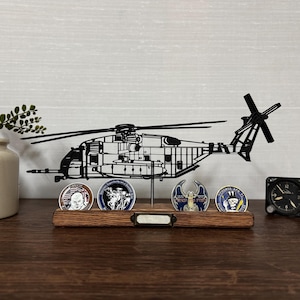 May include: A black metal helicopter silhouette display with a wooden base holding four military challenge coins. A small black clock and a decorative plant in a cream-colored pot are also present.