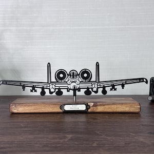 May include: A black metal A-10 Warthog airplane silhouette mounted on a wooden base. The model has detailed wings, engines, and weaponry. A small metal tag reads "A-10 Warthog". A vintage clock is to the right.