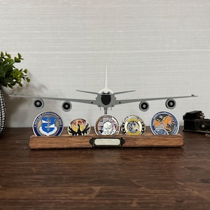 May include: A wooden display stand holding five military challenge coins, with a model airplane in the background. The coins have colorful designs and text. A small plant in a decorative pot is on the left.