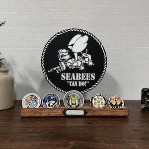 May include: A wooden display stand holding six military challenge coins. A large black circular emblem with the text "SEABEES CAN DO!" is behind the coins. A small clock and a decorative plant in a cream-colored pot are also visible.