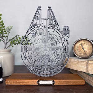 May include: A detailed, silver-colored Millennium Falcon model, laser-cut from metal, displayed on a wooden base. The model is intricately designed, showcasing the ship's iconic features. A small label is visible on the base. Other items include a plant, a gauge, and books.