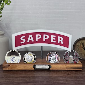 May include: A wooden display stand holding four collectible coins. The stand features a sign that reads "SAPPER" in white letters on a red background. The coins have various designs and colors, including a running figure and a portrait.