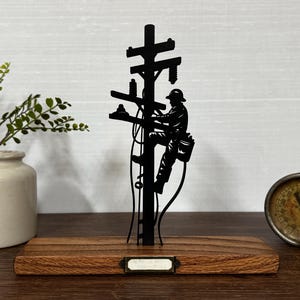 May include: A black metal silhouette of a lineman climbing a utility pole, mounted on a wooden base. The silhouette is detailed, showing the lineman's tools and gear. A small, vintage gauge and a ceramic vase with greenery are also in the image.