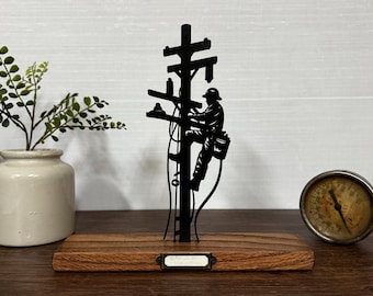 May include: A black metal silhouette of a lineman climbing a utility pole, mounted on a wooden base. The silhouette is detailed, showing the lineman's tools and gear. A small, vintage gauge and a ceramic vase with greenery are also in the image.