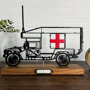 May include: A black metal ambulance truck model with a red cross on the side, mounted on a wooden base. The model has intricate details and a tall antenna. A vintage gauge is visible in the background.