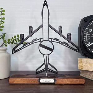 May include: A black metal airplane sculpture on a wooden base, with a circular design in the center. The sculpture is displayed with a black altitude clock and a small plant in a cream-colored pot.