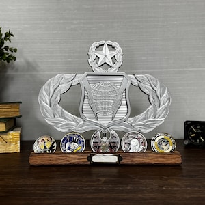 May include: A silver-colored military emblem with a star and globe design, displayed above a wooden base holding several round coins. The coins feature colorful designs and text. A small black clock is to the right.