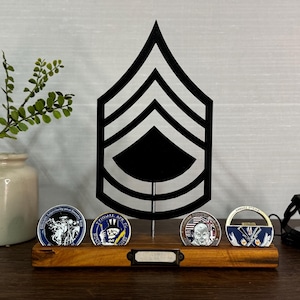 May include: A wooden display stand holding four challenge coins, with a black metal Sergeant Major rank insignia in the background. A small vase with greenery is on the left. The coins have various designs and colors.