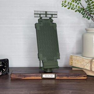 May include: A detailed model of a radar system, predominantly olive green, mounted on a wooden base. The model features a grid-like antenna and a detailed structure. A vintage clock and a decorative vase with greenery are also visible.