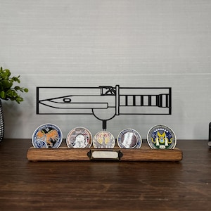 May include: A wooden display stand holding five collectible coins. Above the coins is a black metal silhouette of a bayonet. A small black clock is on the right. A potted plant is on the left.