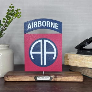 May include: A wooden display featuring a red and blue military emblem with the word "AIRBORNE" at the top. The emblem has a white circle with the letters "AA" inside. A small plaque with text is at the base.