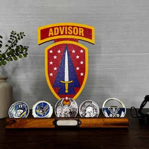 May include: A wooden display stand holding five military challenge coins, with a large emblem reading "ADVISOR" in the background. The coins feature various designs and colors, and the stand is on a dark wooden surface.