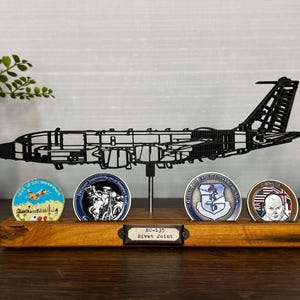 May include: A black metal RC-135 Rivet Joint aircraft model with a wooden base. The base holds four military challenge coins. The model is displayed with a small clock and a decorative plant in a ceramic pot.