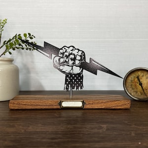 May include: A black metal sculpture of a clenched fist holding a lightning bolt, mounted on a wooden base. The sculpture has a vintage aesthetic, with a small label holder on the base. A small plant in a ceramic pot and a gauge are also in the image.
