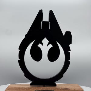 May include: A black Millennium Falcon silhouette with a white Rebel Alliance symbol is mounted on a rectangular wooden base. The base has a small metal tag that reads "YT 492727ZED Millennium Falcon".