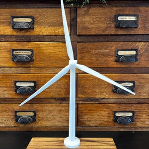 May include: A white miniature wind turbine model with three blades, mounted on a rectangular wooden base. The model is set against a backdrop of a wooden cabinet with multiple drawers. The blades are angled to catch the wind.