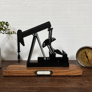 May include: A black metal oil pump jack model on a wooden base with a small metal label. A vintage gauge and a small vase with greenery are also visible. The oil pump jack is a decorative item.