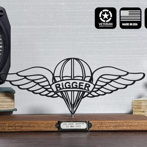 May include: Black metal "Rigger" emblem with wings, parachute, and text on a wooden base. Includes a small nameplate with text. The background features a clock, books, and other decorative items.