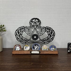 May include: A wooden display stand holding five commemorative coins, with a black metal emblem in the background. A small black clock is to the right. A small plant in a cream-colored vase is to the left.