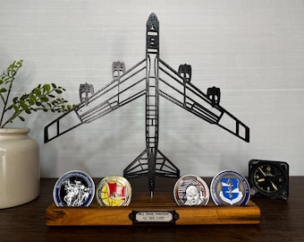 May include: A black 3D-printed B-52 aircraft silhouette mounted on a rectangular wooden base. The base holds four circular military challenge coins and a small metal plaque engraved with "Maj Jack Teacher 01 Oct 1995" in black text.