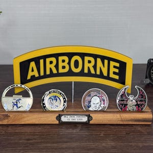May include: A wooden display stand holding four military challenge coins, with an "AIRBORNE" sign in the background. The coins feature various designs and colors. A vintage clock is visible on the right.