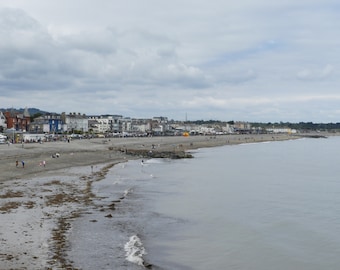 May include: A view of a beach with a town in the background. The beach is covered in sand and there are people walking on it. The town is made up of buildings with white walls and red roofs. The sky is cloudy and gray.