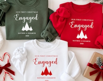 May include: Three crewneck sweatshirts in olive green, red, and white. Each features the text "OUR FIRST CHRISTMAS Engaged" with a graphic of three pine trees and the names "MATTHEW & ELISABETH" followed by the year "2026" printed on the front.