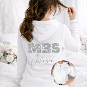 May include: White zip-up hoodie with the word "MRS" in silver glitter lettering on the back, followed by the name "Johnson" in a script font. The date "04.25.2026" is printed on the sleeve. A small image of the hoodie is in the bottom right corner.