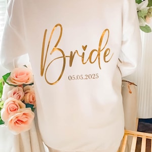 May include: A white sweatshirt with the word "Bride" in gold script across the back, along with the date 05.05.2025. The sweatshirt is surrounded by pink and peach roses, suggesting a bridal theme.