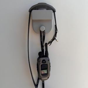 Könnte beinhalten: Ein graues Dogtra-Fern-Trainingshalsband mit einem schwarzen Halsband und einer schwarzen und grauen Fernbedienung. Die Fernbedienung hat einen Bildschirm und Knöpfe. Das Halsband ist mit einem Haken an der Wandhalterung befestigt.