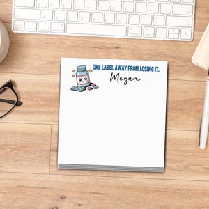 May include: A white notepad with the text "ONE LABEL AWAY FROM LOSING IT." and the name "Megan" in blue script. The notepad features a cartoon illustration of a pill bottle and pills. The notepad is on a wooden desk.