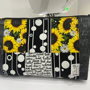 May include: Black and white zippered pouch with a patchwork design featuring sunflowers, white daisies, and circles. The pouch has a yellow tassel and a small fabric label with the text "The Stitchin'  Lady". The pouch has a handwritten message on the front that reads "dream cutest best adorable sweet hello sisters love best brea hello sisters adorat sweet dream adorat ove best cutest swee sisters sweet dream cutest adorable cutest love cutest".
