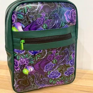 Puede incluir: Una mochila de color verde oscuro con un estampado vibrante de rosas moradas, cuervos negros y siluetas de dragones. La bolsa incluye un bolsillo frontal horizontal con cremallera, herrajes verde lima y un tirador a juego en el compartimento principal.