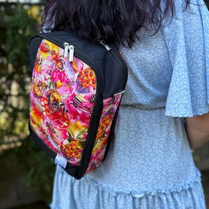 May include: Black backpack with a colourful floral and bee print. The design features pink, yellow, and orange flowers with honeycombs and bees. The backpack has a black zip and straps, and is worn over the shoulder.