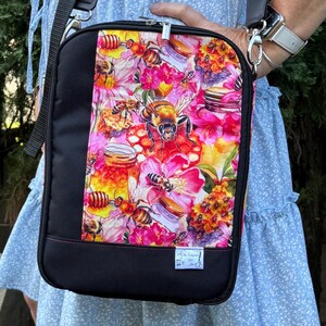 May include: A black crossbody bag with a zip closure. The front of the bag features a colourful print of bees, flowers, and honey jars. The bag has a black strap and a small tag that says "The Scrappy Stitcher".