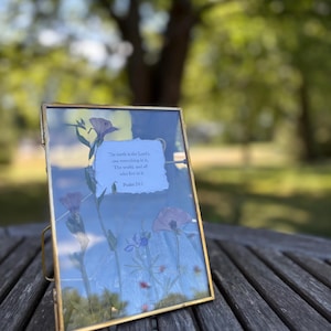 May include: A rectangular gold-framed glass display featuring pressed flowers and the text "The earth is the Lord's, and everything in it, The world, and all who live in it Psalm 24:1". The display is on a wooden table.