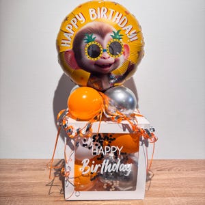 Könnte beinhalten: Eine weiße quadratische Box mit einem Sichtfenster, das den Text Happy Birthday zeigt, gefüllt mit orangefarbenen, silbernen und schwarzen Luftballons. Ein runder gelber Folienballon mit einem Affen mit Ananas-Sonnenbrille und dem Text Happy Birthday befindet sich oben.