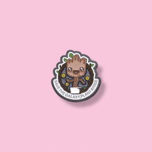 May include: A black and white enamel pin featuring a cartoon character of Groot from Guardians of the Galaxy. The character is holding a pot and has a green leafy crown. The text around the character reads "Save the Galaxy, Plant a Tree".