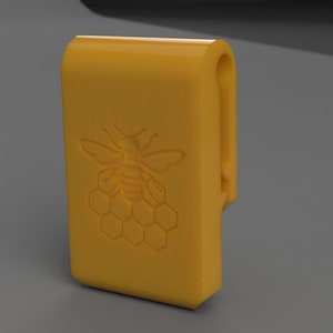 May include: A yellow rectangular clip with a curved top and a bee and honeycomb design. The clip is made of a solid material and is set against a gray background. The bee is centered on the clip.